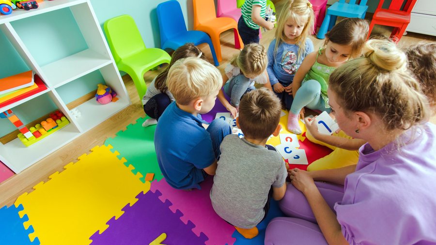 Erzieherin sitzt mit Kindern im Kreis auf bunten Spielmatten